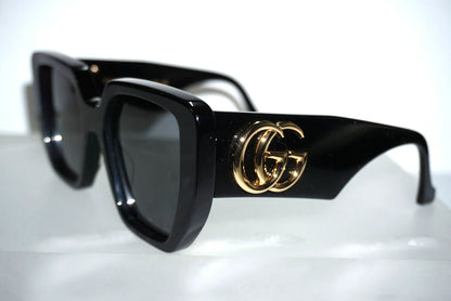 Black square sunglasses with a gold interlocking GG logo on the temple, displayed on a reflective white surface.