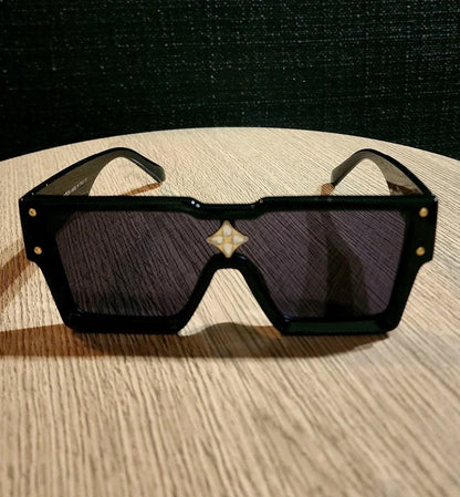 A pair of oversized square sunglasses with a glossy black frame, dark tinted lenses, and a small white geometric logo centered above the lenses, resting on a light wood surface.