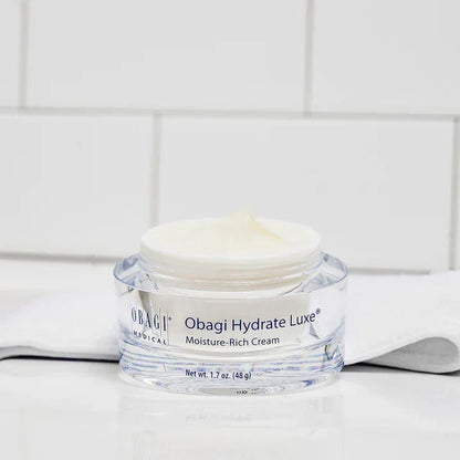 A clear plastic jar of Obagi Magical Obagi Hydrate Luxe Moisture-Rich Cream with white cream inside, set on a white surface with a white tiled background.