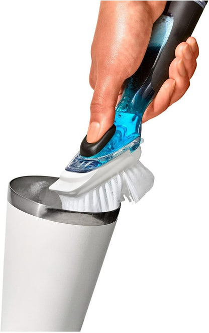 A hand holding a blue and black handheld cleaning brush above a white cylindrical container with a metal rim, shown against a plain white background.