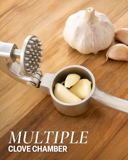 A stainless steel garlic press with a textured clove chamber and handle, shown with garlic cloves inside and whole garlic bulbs on a wooden surface. Text overlay reads 'MULTIPLE CLOVE CHAMBER'.