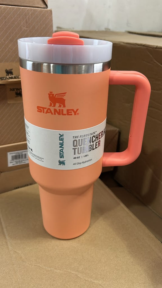 A Stanley QuenchMaster travel tumbler in a peach-orange color, featuring a white lid with a coral accent, a stainless steel rim, and a white product label on a cardboard background.