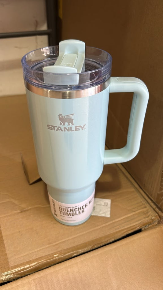 A light blue Stanley Quencher 32 oz handled tumbler with a clear lid and white flip-top spout, resting on a cardboard box.