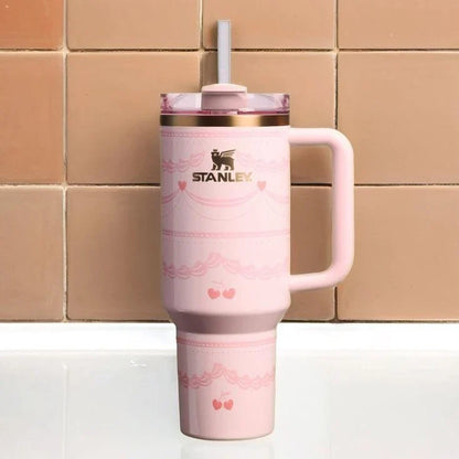 A pink Stanley travel mug with a handle, a clear lid with a white straw, and rose gold trim, set against a background of tan ceramic tiles.