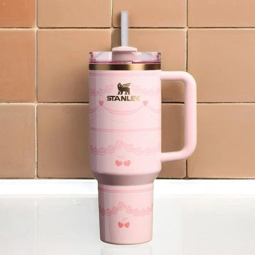 A pink Stanley travel mug with a handle, a clear lid with a white straw, and rose gold trim, set against a background of tan ceramic tiles.