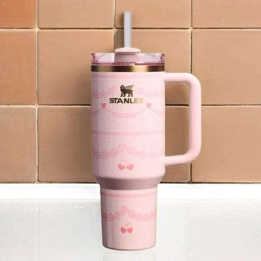 A pink Stanley travel mug with a handle, a clear lid with a white straw, and rose gold trim, set against a background of tan ceramic tiles.