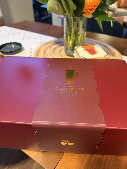 A red rectangular gift box with a scalloped flap closure and gold printed text reading 'Guatemalan H2.0 Tumbler Hot Tea' sits on a wooden table.