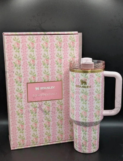 A pink floral Stanley tumbler with a white handle and lid, shown next to its matching pink patterned gift box on a dark background.