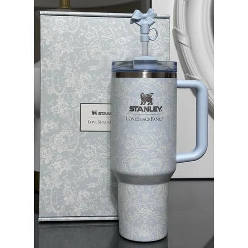 A light blue Stanley Love Shack Fancy insulated tumbler with a matching lid and handle, shown in front of its patterned gift box on a dark surface.