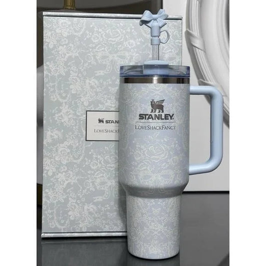 A light blue Stanley Love Shack Fancy insulated tumbler with a matching lid and handle, shown in front of its patterned gift box on a dark surface.